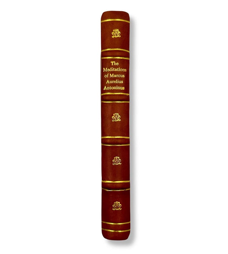 May include: A hardcover book with a dark brown leather cover. The spine features gold lettering that reads "The Meditations of Marcus Aurelius Antoninus." Gold bands and decorative emblems adorn the spine.