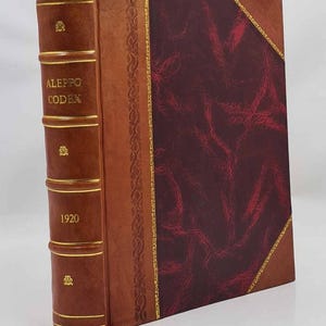 Peut inclure: Un livre relié en cuir avec une tranche et une couverture marron. La tranche porte l'inscription "ALEPPO CODEX 1920" en lettres dorées. La couverture présente un motif bordeaux avec des bordures dorées et un signet rouge.