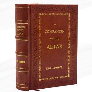 Può includere: Un libro con copertina rigida intitolato "A Companion to the Altar" di Tho. Comber. Il libro ha una copertina marrone rossiccio scuro con scritte dorate e bordi dorati decorativi. Anche il dorso presenta scritte dorate.