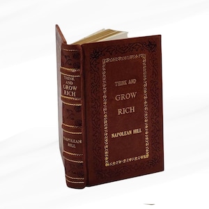 May include: A brown hardcover book titled "Think and Grow Rich" by Napoleon Hill. The cover and spine feature gold lettering and decorative detailing. The book's title and author are clearly visible.