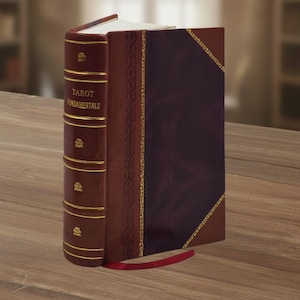 Puede incluir: Un libro de tapa dura titulado "TAROT FUNDAMENTALS" con una cubierta y lomo de color burdeos. El lomo está adornado con detalles dorados y elementos decorativos. Una cinta roja sirve como marcador.
