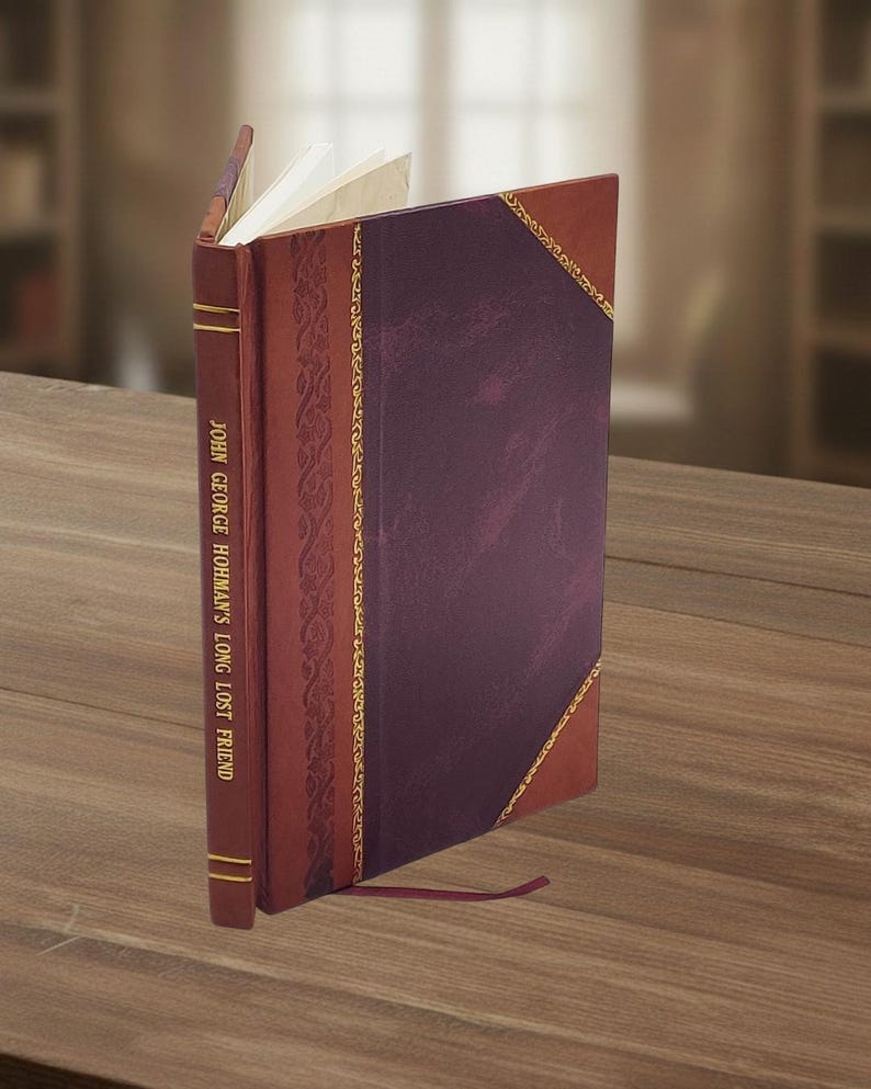 May include: A hardcover book with the title "JOHN GEORGE HOHMAN'S LONG LOST FRIEND" in gold lettering. The book has a burgundy cover with a dark purple center panel, accented with gold trim. A burgundy ribbon bookmark is visible.