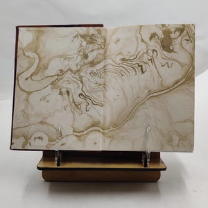 Puede incluir: Un libro abierto con p&aacute;ginas veteadas, expuesto sobre un atril de madera. Las p&aacute;ginas presentan un patr&oacute;n arremolinado en tonos beige y dorado. La cubierta del libro es de color burdeos oscuro.