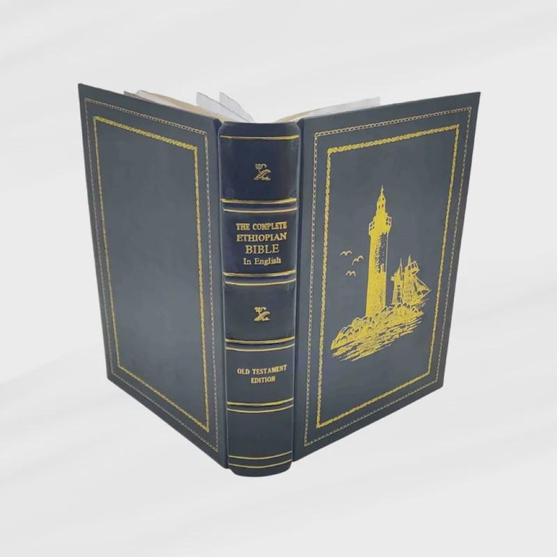 K&ouml;nnte beinhalten: Ein offenes Hardcover-Buch mit dem Titel "The Complete Ethiopian Bible In English" mit marineblauem Einband und goldenen Akzenten. Das Cover zeigt eine goldene Illustration eines Leuchtturms und eines Schiffes.