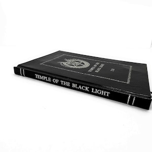 K&ouml;nnte beinhalten: Ein schwarzes Buch mit dem Titel "TEMPLE OF THE BLACK LIGHT" in wei&szlig;er Schrift. Das Cover zeigt ein silbernes Pentagramm-Design in einem Kreis. Der Buchr&uuml;cken hat wei&szlig;e N&auml;hte und ein schwarzes Lesezeichen. Das Buch liegt auf einer wei&szlig;en Oberfl&auml;che.