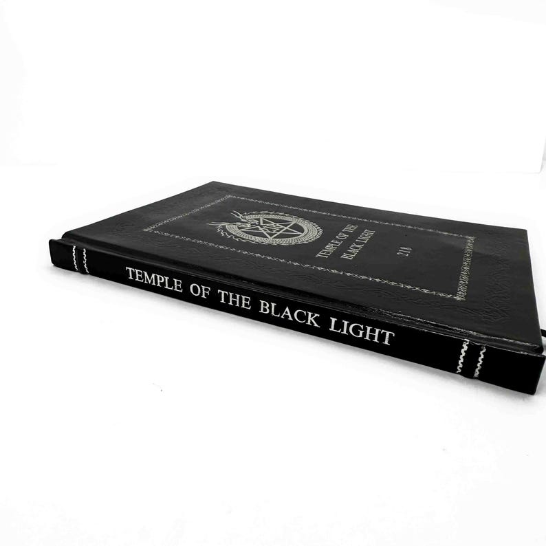 K&ouml;nnte beinhalten: Ein schwarzes Buch mit dem Titel "TEMPLE OF THE BLACK LIGHT" in wei&szlig;er Schrift. Das Cover zeigt ein silbernes Pentagramm-Design in einem Kreis. Der Buchr&uuml;cken hat wei&szlig;e N&auml;hte und ein schwarzes Lesezeichen. Das Buch liegt auf einer wei&szlig;en Oberfl&auml;che.