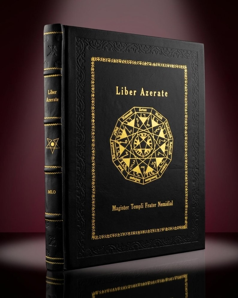 May include: A black book titled "Liber Azerate" with gold lettering and a gold geometric design. The book's spine has gold accents and the text "Liber Azerate" and "MLO". The book is on a reflective surface.