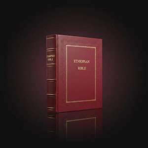 Op de afbeelding: Een rood leren boek met de titel "Ethiopian Bible" met gouden letters en randen. De rug heeft gouden letters en decoratieve elementen. Het boek staat rechtop op een reflecterend oppervlak, met een donkere achtergrond.