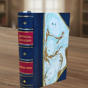 Op de afbeelding: Een gebonden boek met de titel Mythical Monsters van Charles Gould. De rug is donkerblauw met gouden accenten en twee rode panelen met gouden tekst. De kaft heeft een gemarmerd patroon in lichtblauw, wit en goud, omlijst door donkerblauw.