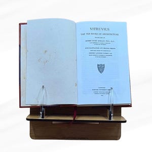 May include: An open book titled "VITRUVIUS The Ten Books on Architecture" displayed on a wooden book stand. The book has a red cover and is propped up by clear acrylic supports. The text is in black font on a light blue background.