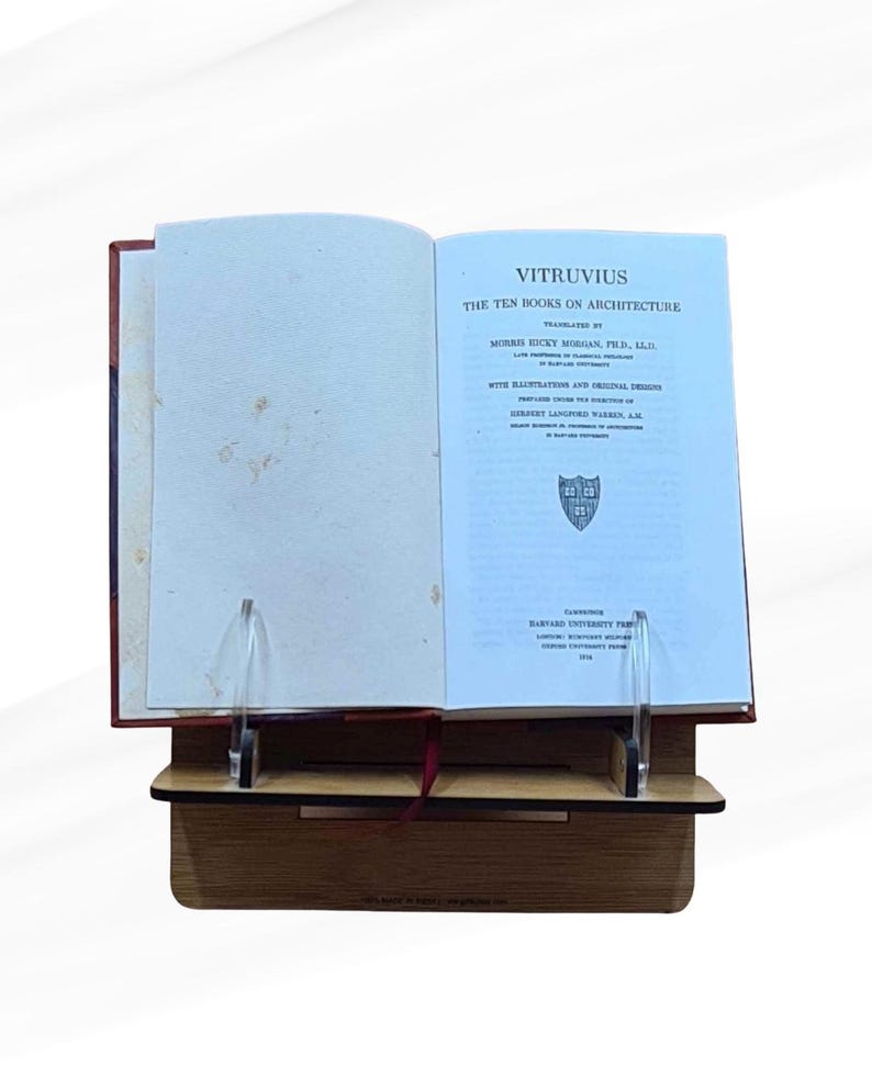May include: An open book titled "VITRUVIUS The Ten Books on Architecture" displayed on a wooden book stand. The book has a red cover and is propped up by clear acrylic supports. The text is in black font on a light blue background.