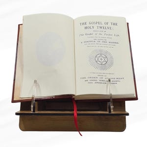 May include: An open book titled "The Gospel of the Holy Twelve" with a red cover and a red ribbon bookmark. The book is displayed on a wooden stand with clear acrylic supports. The text is in black ink on aged paper.