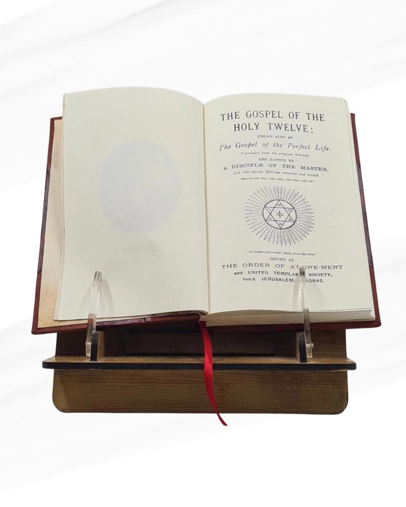 May include: An open book titled "The Gospel of the Holy Twelve" with a red cover and a red ribbon bookmark. The book is displayed on a wooden stand with clear acrylic supports. The text is in black ink on aged paper.