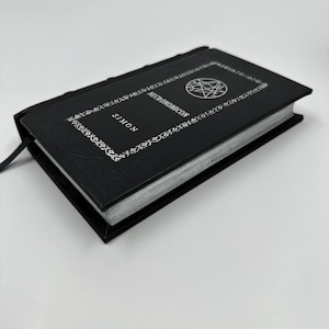 Puede incluir: Un libro de tapa dura negro titulado Necronomicon de Simon, que presenta un sello circular plateado en la portada. La cubierta est&aacute; decorada con un patr&oacute;n de borde floral blanco y los bordes de las p&aacute;ginas tienen un acabado en color plata met&aacute;lico.