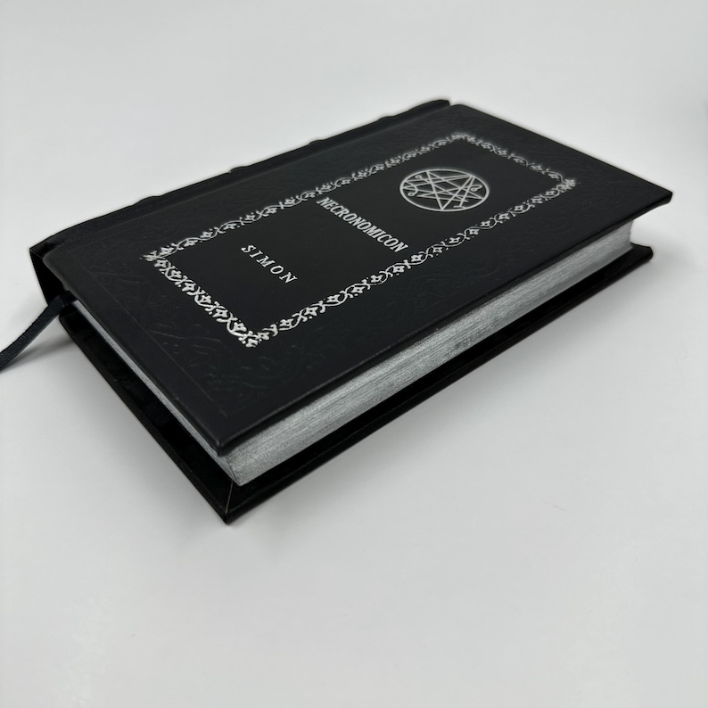 Puede incluir: Un libro de tapa dura negro titulado Necronomicon de Simon, que presenta un sello circular plateado en la portada. La cubierta est&aacute; decorada con un patr&oacute;n de borde floral blanco y los bordes de las p&aacute;ginas tienen un acabado en color plata met&aacute;lico.