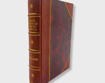 Op de afbeelding: Een hardcover boek getiteld "Honey Plants of North America" met een rood lint-bladwijzer. Het boek heeft een bruine leren kaft met gouden letters en accenten. De kaft heeft een getextureerd, bordeauxrood paneel.