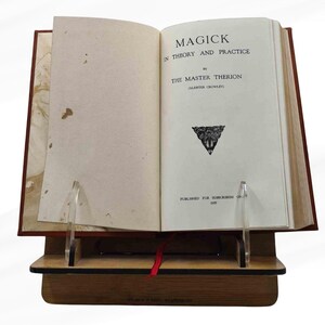 Peut inclure: Livre ouvert intitul&eacute; &laquo; MAGICK IN THEORY AND PRACTICE &raquo; par The Master Therion (Aleister Crowley). Le livre est pr&eacute;sent&eacute; sur un pr&eacute;sentoir en bois avec des supports en acrylique transparents. Un ruban rouge marque une page.