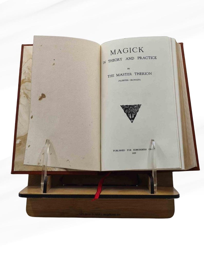 Peut inclure: Livre ouvert intitul&eacute; &laquo; MAGICK IN THEORY AND PRACTICE &raquo; par The Master Therion (Aleister Crowley). Le livre est pr&eacute;sent&eacute; sur un pr&eacute;sentoir en bois avec des supports en acrylique transparents. Un ruban rouge marque une page.
