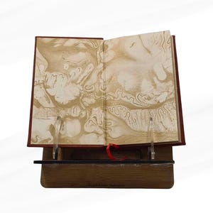Pode incluir: Um livro aberto com p&aacute;ginas marmoreadas, exposto em um suporte de madeira. O livro tem uma capa vermelha e uma fita vermelha como marcador. As p&aacute;ginas apresentam um design abstrato e rodopiante em tons de ouro e creme.