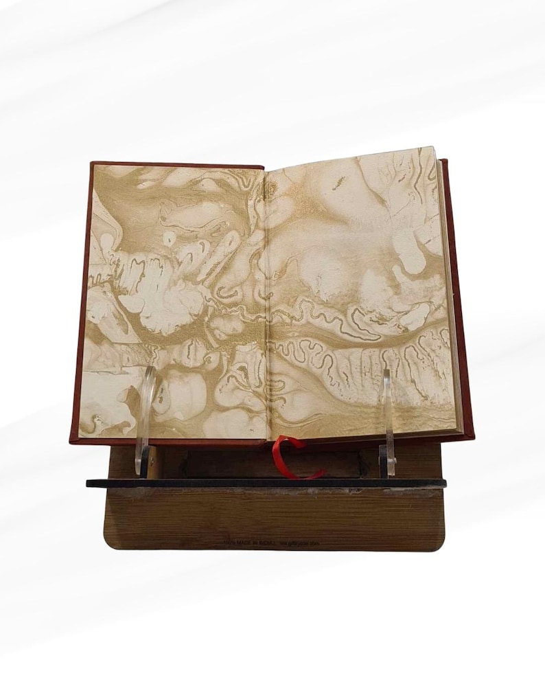 Pode incluir: Um livro aberto com p&aacute;ginas marmoreadas, exposto em um suporte de madeira. O livro tem uma capa vermelha e uma fita vermelha como marcador. As p&aacute;ginas apresentam um design abstrato e rodopiante em tons de ouro e creme.
