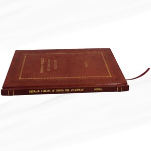 Puede incluir: Un libro de tapa dura titulado "Emerald Tablets of Thoth the Atlantean" con letras y adornos dorados. El libro est&aacute; encuadernado en un material similar al cuero marr&oacute;n rojizo y tiene una cinta roja como marcador. El nombre del autor, DOREAL, est&aacute; impreso en el lomo.