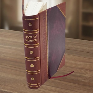 May include: A hardback book with a brown leather spine and cover. The spine features the title "BOOK OF MORMON" in gold lettering, with decorative gold accents. A red ribbon bookmark extends from the book's edge. The book is open, revealing white pages.