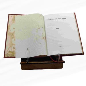 Puede incluir: Un libro abierto titulado "Emerald Tablets Of Thoth The Atlantean" con el a&ntilde;o 1930 visible. El libro est&aacute; apoyado en un soporte de madera. La portada es de color rojo oscuro.