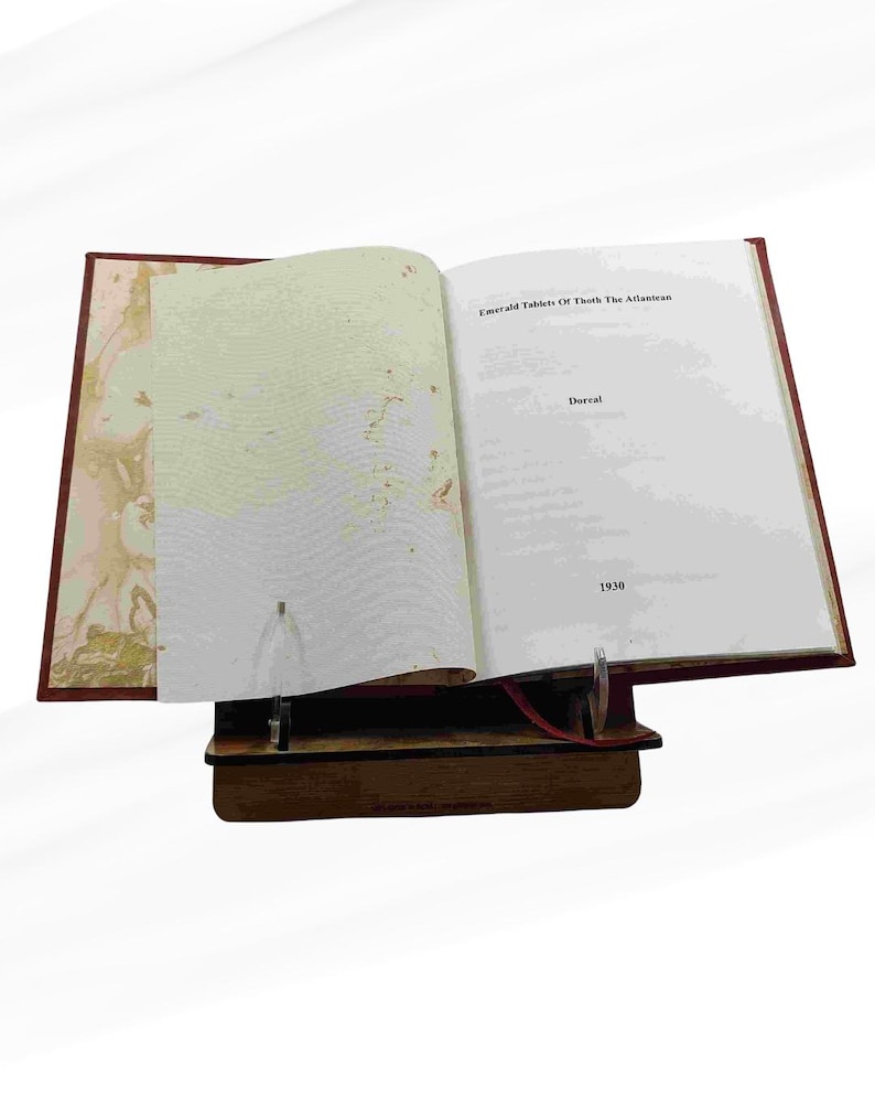 Puede incluir: Un libro abierto titulado "Emerald Tablets Of Thoth The Atlantean" con el a&ntilde;o 1930 visible. El libro est&aacute; apoyado en un soporte de madera. La portada es de color rojo oscuro.
