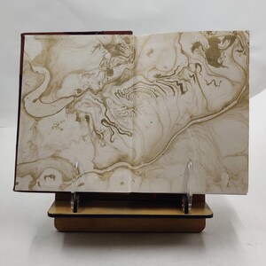 May include: An open book with marbled pages, displayed on a wooden book stand. The pages feature a swirling pattern in shades of beige and gold. The book's cover is a deep burgundy color.