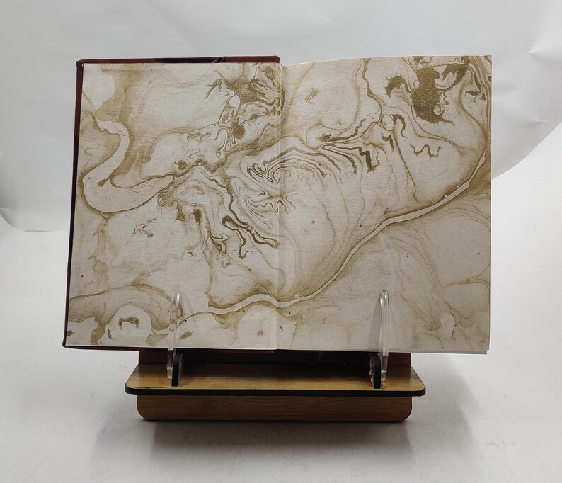 May include: An open book with marbled pages, displayed on a wooden book stand. The pages feature a swirling pattern in shades of beige and gold. The book's cover is a deep burgundy color.