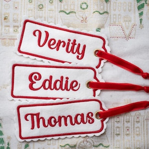 May include: Three rectangular white felt gift tags with scalloped edges and red embroidered borders. Each tag features a red embroidered name: Verity, Eddie, or Thomas. A gold metal eyelet secures a thin red ribbon to each individual tag.