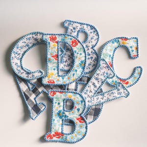 May include: A collection of fabric letters including A, B, C, and D, featuring floral patterns in red, yellow, and blue, alongside black and white buffalo check patterns. Each letter is finished with a blue stitched border around the edges.
