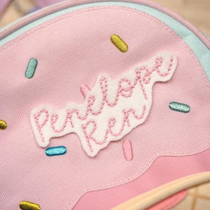 Op de afbeelding: Roze rugzak met een grillig ontwerp, met een witte patch geborduurd met "Penelope Ren" in roze draad. De tas is versierd met kleurrijke geborduurde sprinkles en een lichtblauwe rand.