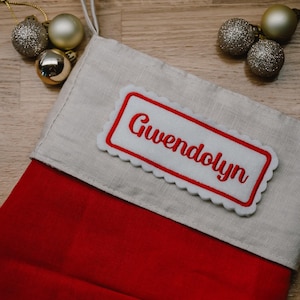 May include: A festive Christmas stocking, red with a light beige cuff, personalized with the name "Gwendolyn" in red embroidery on a white patch. Glittering gold and silver baubles are arranged nearby.