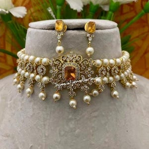 Puede incluir: Un conjunto de collar y pendientes dorados. El collar presenta una piedra central cuadrada de color &aacute;mbar, rodeada de piedras claras m&aacute;s peque&ntilde;as y perlas colgantes. Los pendientes tienen piedras &aacute;mbar a juego y gotas de perlas. El texto "special jeweller india" es visible.