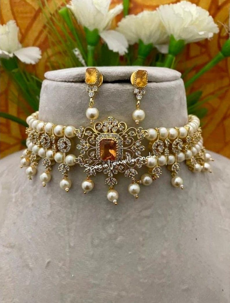 Puede incluir: Un conjunto de collar y pendientes dorados. El collar presenta una piedra central cuadrada de color &aacute;mbar, rodeada de piedras claras m&aacute;s peque&ntilde;as y perlas colgantes. Los pendientes tienen piedras &aacute;mbar a juego y gotas de perlas. El texto "special jeweller india" es visible.