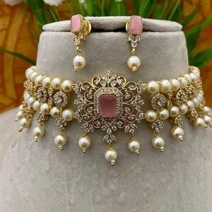 Puede incluir: Un conjunto de collar y pendientes dorados con una piedra rosa en el centro, perlas de imitaci&oacute;n y cristales transparentes. El collar tiene un dise&ntilde;o floral con detalles de perlas colgantes. Los pendientes tienen piedras rosas a juego y gotas de perlas.