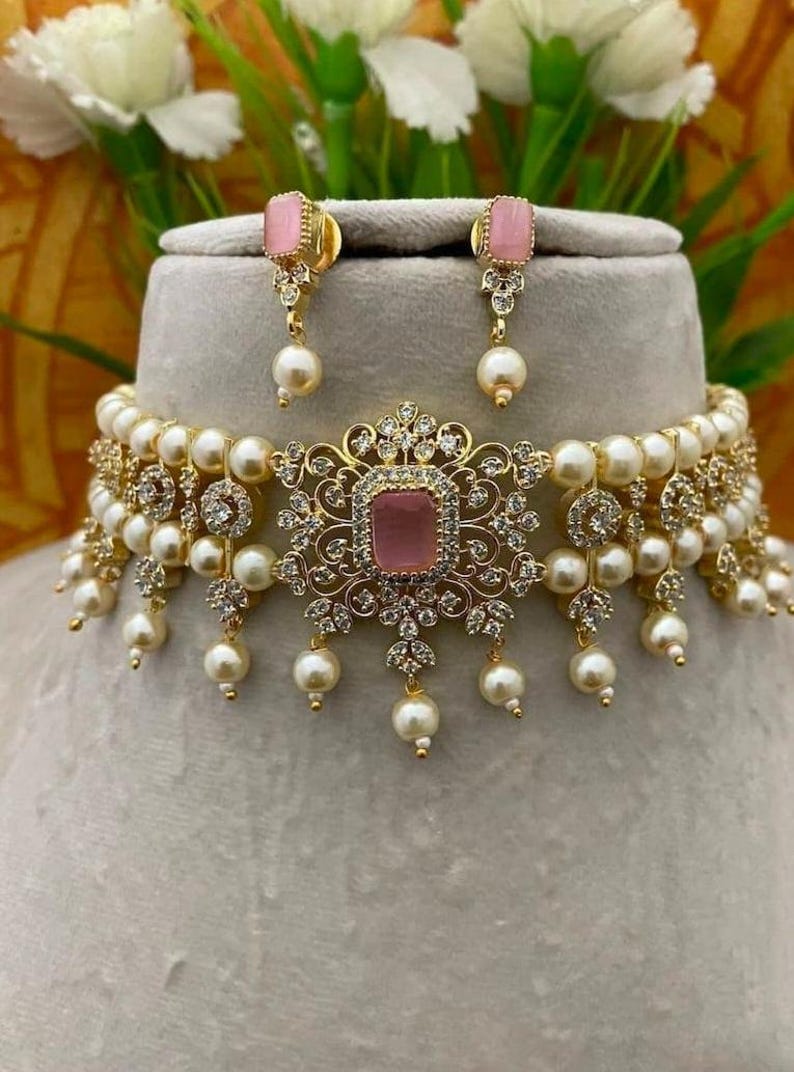 Puede incluir: Un conjunto de collar y pendientes dorados con una piedra rosa en el centro, perlas de imitaci&oacute;n y cristales transparentes. El collar tiene un dise&ntilde;o floral con detalles de perlas colgantes. Los pendientes tienen piedras rosas a juego y gotas de perlas.