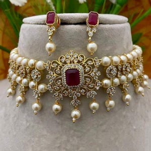 Puede incluir: Un conjunto de collar y pendientes dorados con una piedra central cuadrada de color rub&iacute;, rodeada de filigranas y peque&ntilde;os cristales transparentes. El collar y los pendientes tienen perlas falsas colgantes.