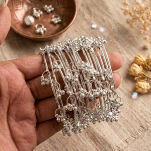 Könnte beinhalten: Eine Hand hält eine Sammlung silberner Armreifen mit baumelnden Anhängern. Die Armreifen haben ein filigranes Design mit kleinen, runden Verzierungen. Eine kleine Kupferschale mit Silberschmuck befindet sich im Hintergrund, zusammen mit Trockenblumen.