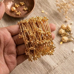 Könnte beinhalten: Eine Hand hält einen Stapel goldfarbener Armreifen. Jeder Armreif ist mit kleinen, hängenden Goldperlen verziert. Eine kleine Kupferschale und Trockenblumen befinden sich im Hintergrund, was auf eine Schmuckpräsentation hindeutet.