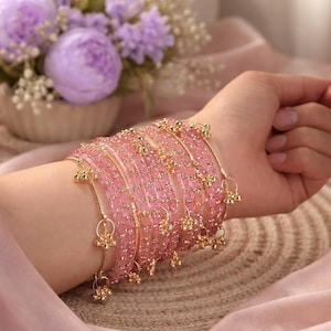 Pode incluir: Pulseiras de vidro rosa com detalhes dourados e pingentes pendurados. As pulseiras são empilhadas em um pulso, com um tecido rosa macio ao fundo. Um buquê de flores roxas também é visível.