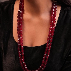 Puede incluir: Un collar de tres hilos compuesto por cuentas ovaladas de color rojo intenso. El collar se lleva alrededor del cuello, con las cuentas cayendo hacia abajo. El fondo es de un color oscuro y sólido, que resalta el rico color y diseño del collar.
