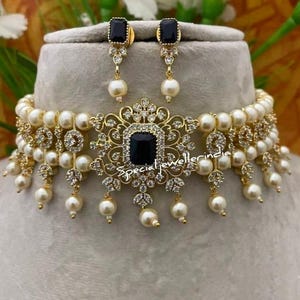 Puede incluir: Juego de collar y pendientes dorados con una piedra cuadrada negra central, rodeada de cristales transparentes y perlas de imitaci&oacute;n colgantes. El collar est&aacute; adornado con m&uacute;ltiples perlas de imitaci&oacute;n. Los pendientes tienen un dise&ntilde;o a juego. El texto "special jewellerindia" es visible.