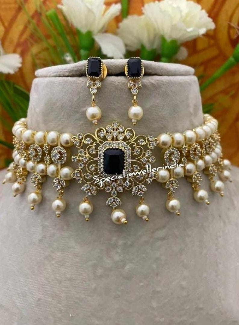 Puede incluir: Juego de collar y pendientes dorados con una piedra cuadrada negra central, rodeada de cristales transparentes y perlas de imitaci&oacute;n colgantes. El collar est&aacute; adornado con m&uacute;ltiples perlas de imitaci&oacute;n. Los pendientes tienen un dise&ntilde;o a juego. El texto "special jewellerindia" es visible.