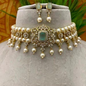 Puede incluir: Un collar dorado y un conjunto de pendientes a juego. El collar presenta una piedra central cuadrada de color verde claro, rodeada por un dise&ntilde;o de filigrana dorada decorativa, acentuada con peque&ntilde;as piedras transparentes y perlas colgantes. Los pendientes tienen un dise&ntilde;o similar.