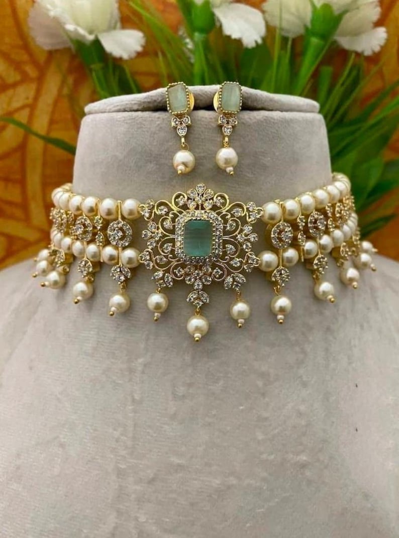 Puede incluir: Un collar dorado y un conjunto de pendientes a juego. El collar presenta una piedra central cuadrada de color verde claro, rodeada por un dise&ntilde;o de filigrana dorada decorativa, acentuada con peque&ntilde;as piedras transparentes y perlas colgantes. Los pendientes tienen un dise&ntilde;o similar.