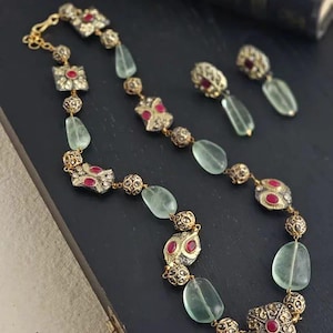 May include: A gold-toned necklace and matching earrings set. The necklace features alternating green, oval-shaped stones and ornate, square and round gold-toned elements with red accents. The earrings have a similar design, with a green stone and a gold-toned top.