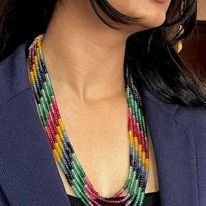 May include: A multi-strand necklace with a variety of colourful gemstones. The necklace displays a vibrant array of colours, including red, green, yellow, and blue. The gemstones are strung together, creating a layered effect. The necklace is worn over a black top and a blue blazer.