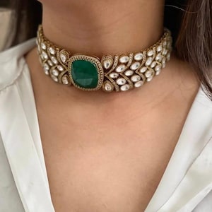 Może przedstawiać: Złoty naszyjnik typu choker z dużym, kwadratowym, szmaragdowozielonym kamieniem szlachetnym pośrodku. Naszyjnik jest ozdobiony licznymi, przezroczystymi, błyszczącymi kamieniami szlachetnymi we wzór kwiatowy, tworząc luksusowy i elegancki design. Naszyjnik jest noszony na szyi.